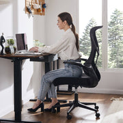 Woman sitting at a desk using a computer in a bright room with large windows.